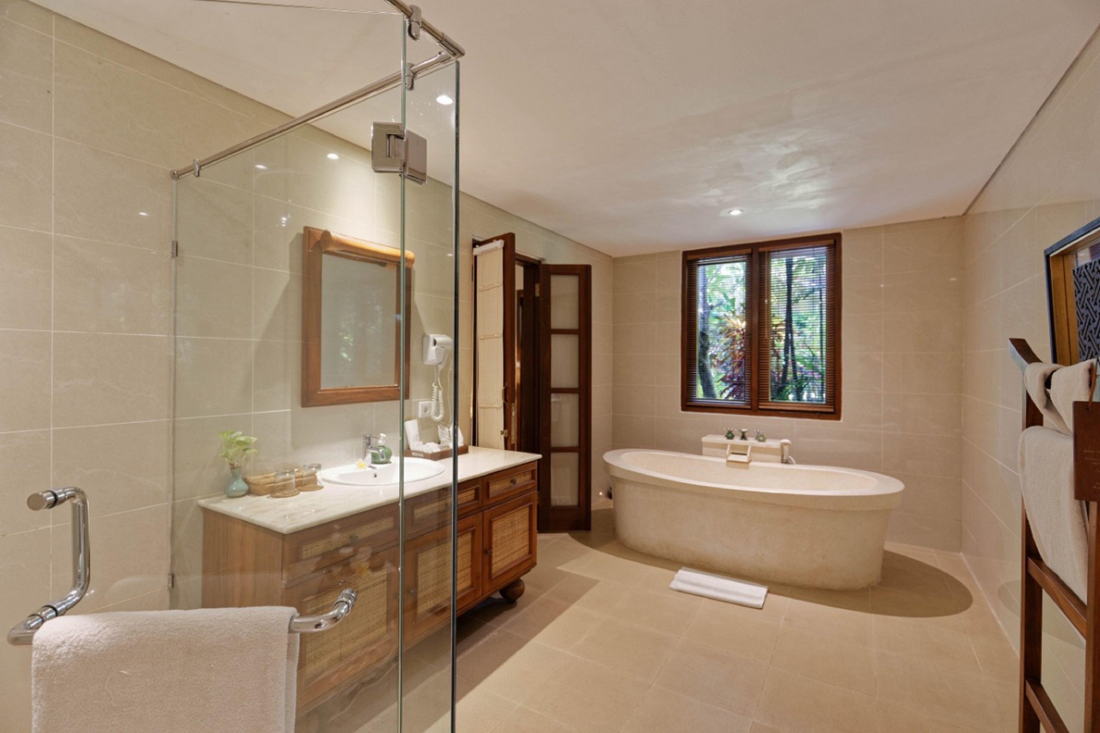 Ein modernes Badezimmer mit einer Glasdusche, einer freistehenden Badewanne, einem Waschtisch mit Spiegel und einem Fenster mit Blick auf Grünflächen.