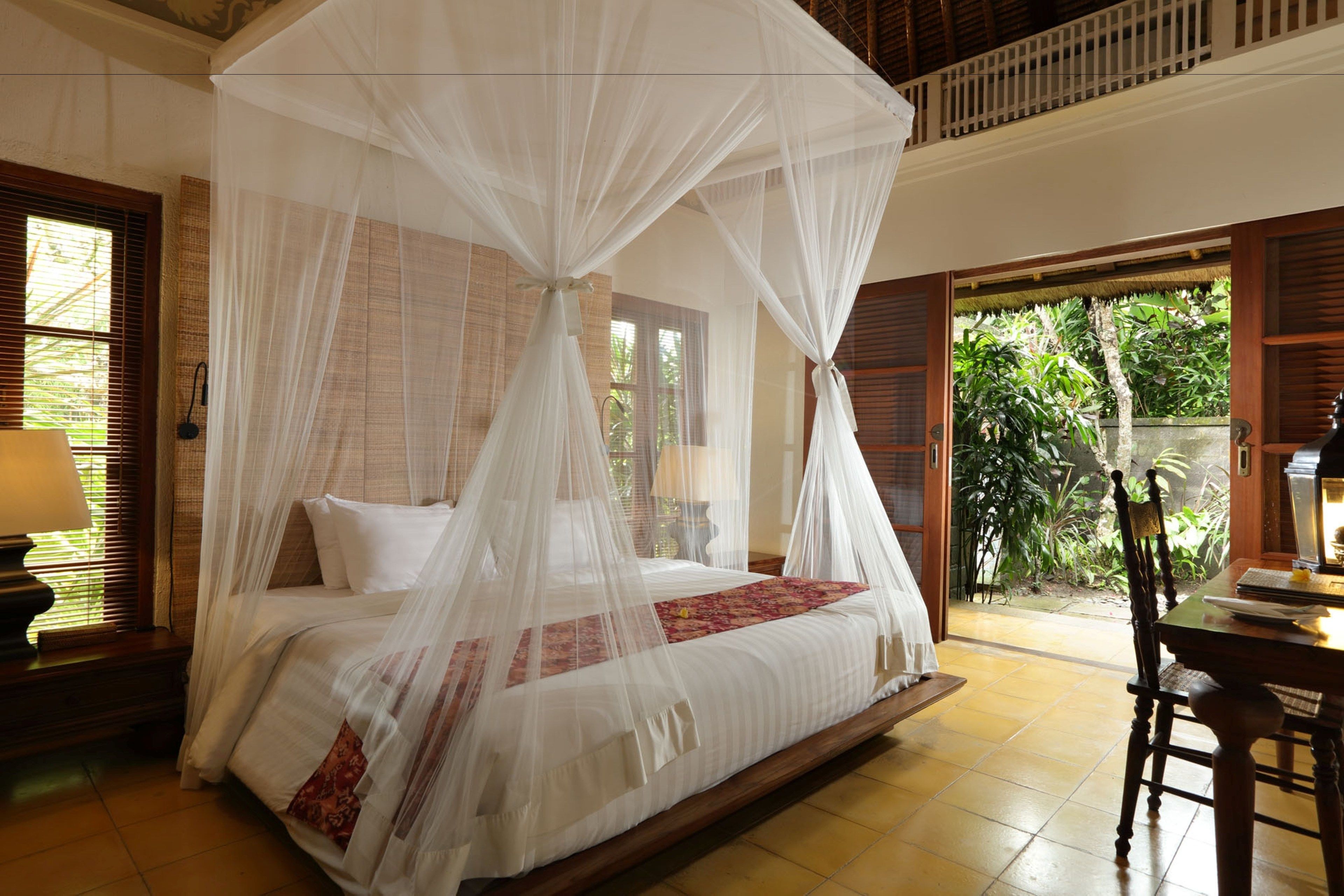 A room with a four-poster bed, a wooden table, and a view of greenery outside.