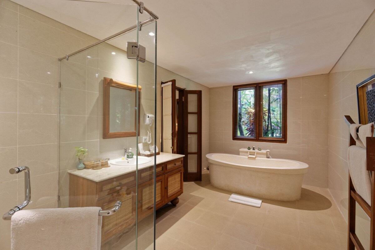A modern bathroom with a glass shower enclosure, a freestanding bathtub, a vanity with a mirror, and a window with a view of greenery.