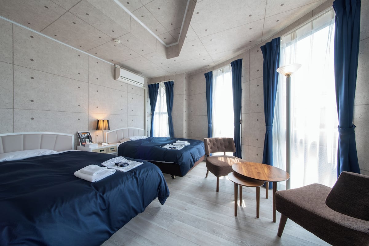 Una habitación de hotel con dos camas, una mesita, una silla y un ventanal con cortinas.