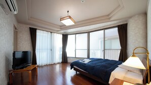 Habitación de hotel con cama, televisor en un mueble bajo, mesita de noche con lámpara y ventanas amplias con cortinas.