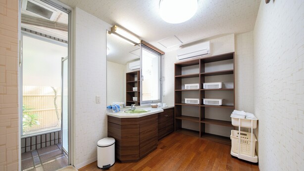 A bathroom with a wooden vanity, a mirror, a sink, and a toilet. There are shelves with baskets and a laundry basket. - Asuka Hotel (Karatsu)
