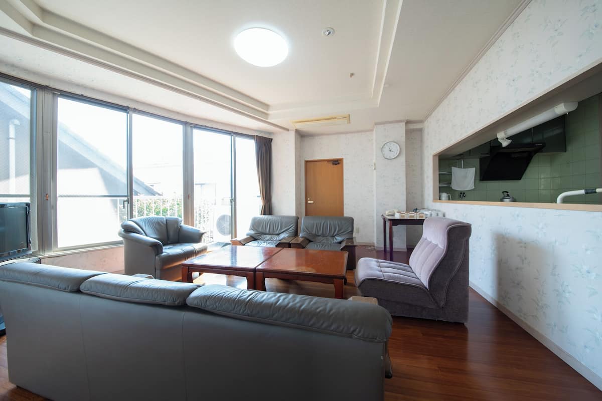 Amplia sala de estar con un sofá grande, dos sillones, una mesa de centro y una mesa de comedor. Hay una zona de cocina con un fregadero y un reloj en la pared.