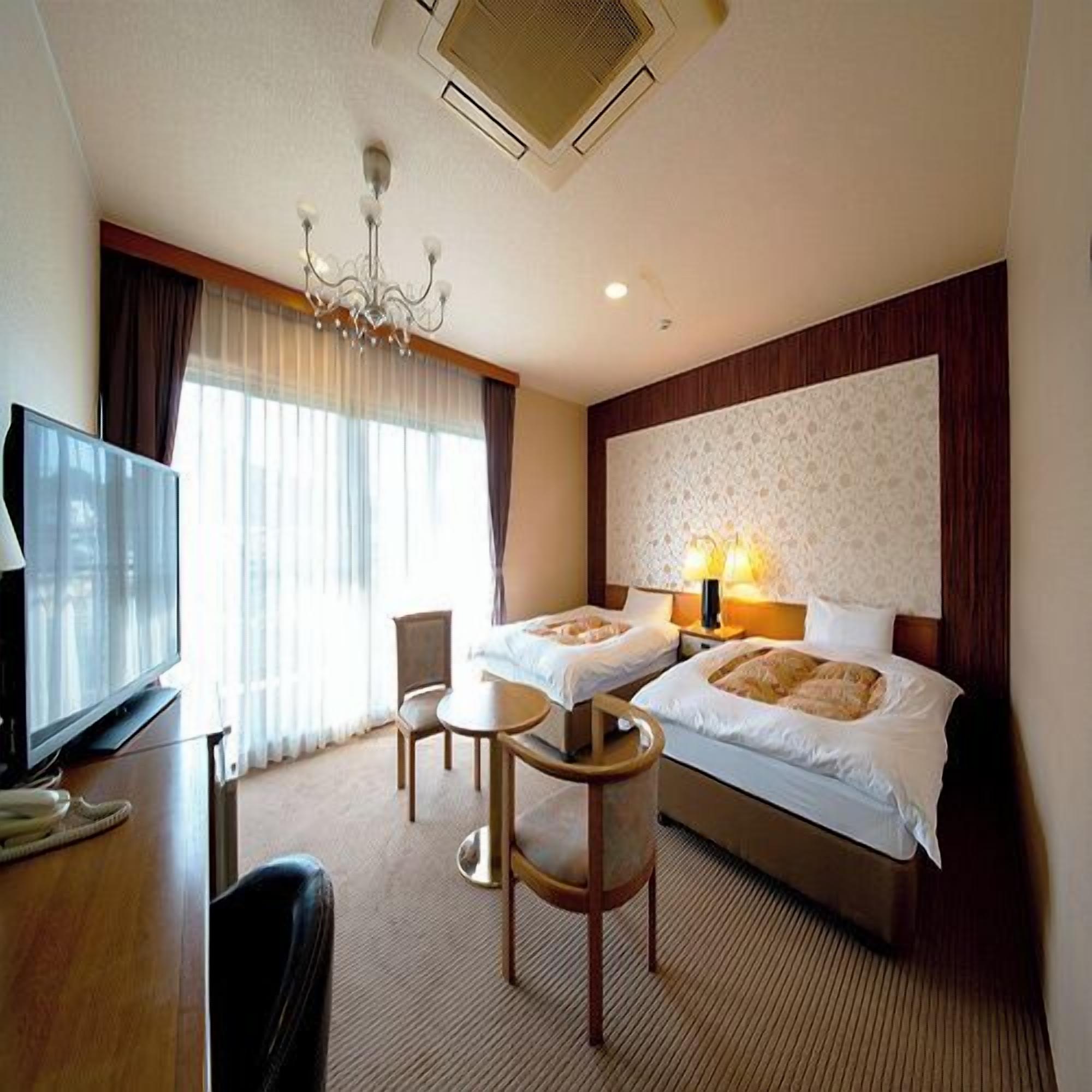 Quarto de hotel com duas camas, televisão, candelabro e uma janela grande com cortinas.