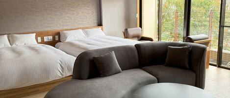 A modern hotel room with a large bed, a sofa, a round table, and a view of trees through large windows.