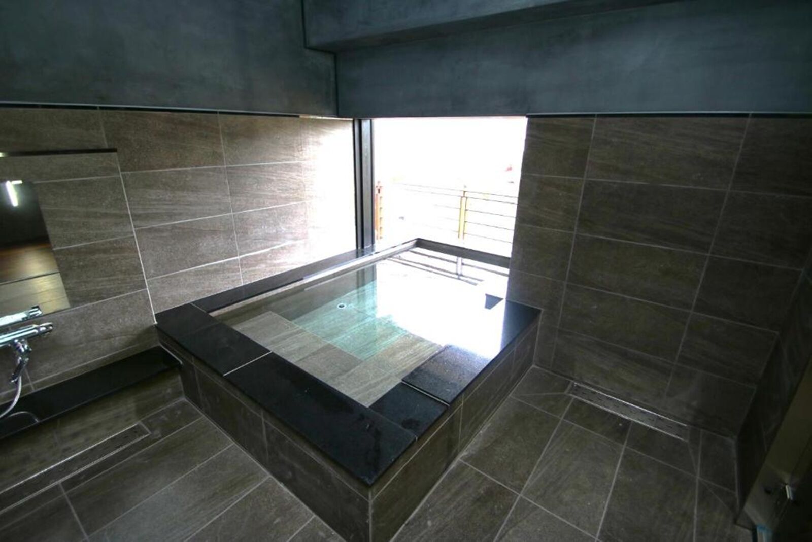 Un baño moderno con bañera de hidromasaje con cerramiento de vidrio, paredes de azulejos oscuros y un área exterior visible a través de la ventana.