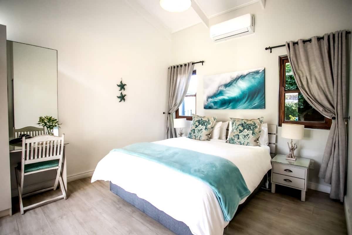 A bedroom with a bed, a desk, a chair, a nightstand, a lamp, and a window with curtains.