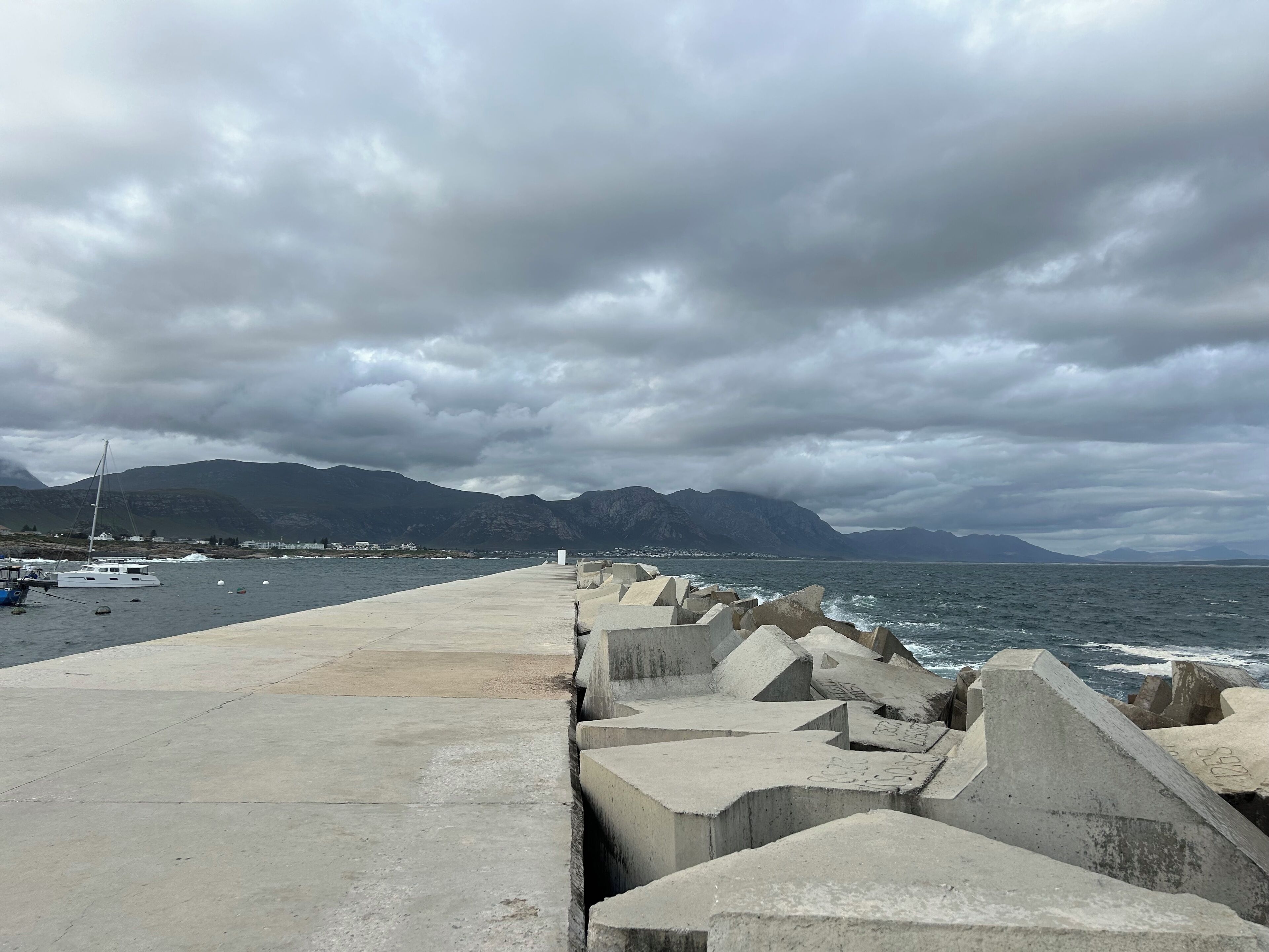 Um cais de concreto se estende sobre a água, com montanhas ao fundo e céu nublado.