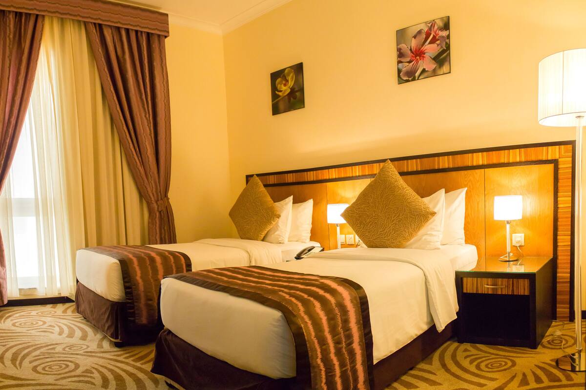 Une chambre d’hôtel avec deux lits, une grande fenêtre avec des rideaux, une table de chevet avec une lampe et deux tableaux accrochés au mur.