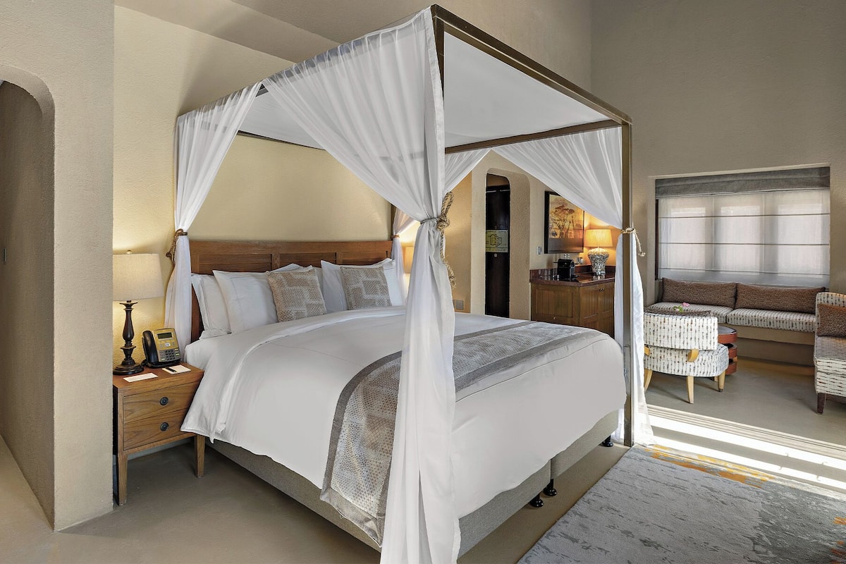 Una cama con dosel y soporte de cuatro postes, con cabecera de madera, una mesita de noche con lámpara y un área de descanso con sofá y sillones.
