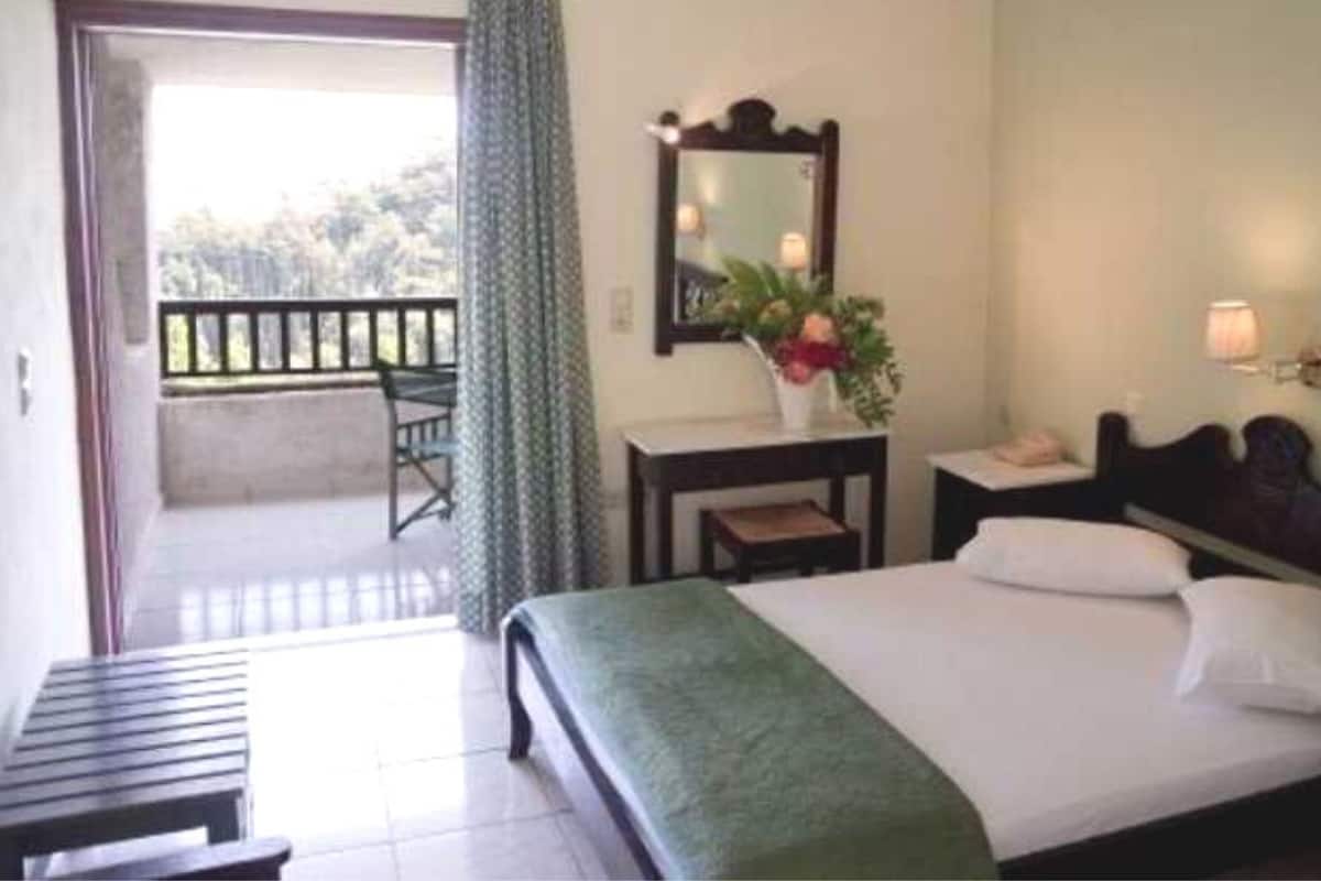 A hotel room with a bed, a desk, a chair, a small table, a mirror, and a view of the outside balcony.