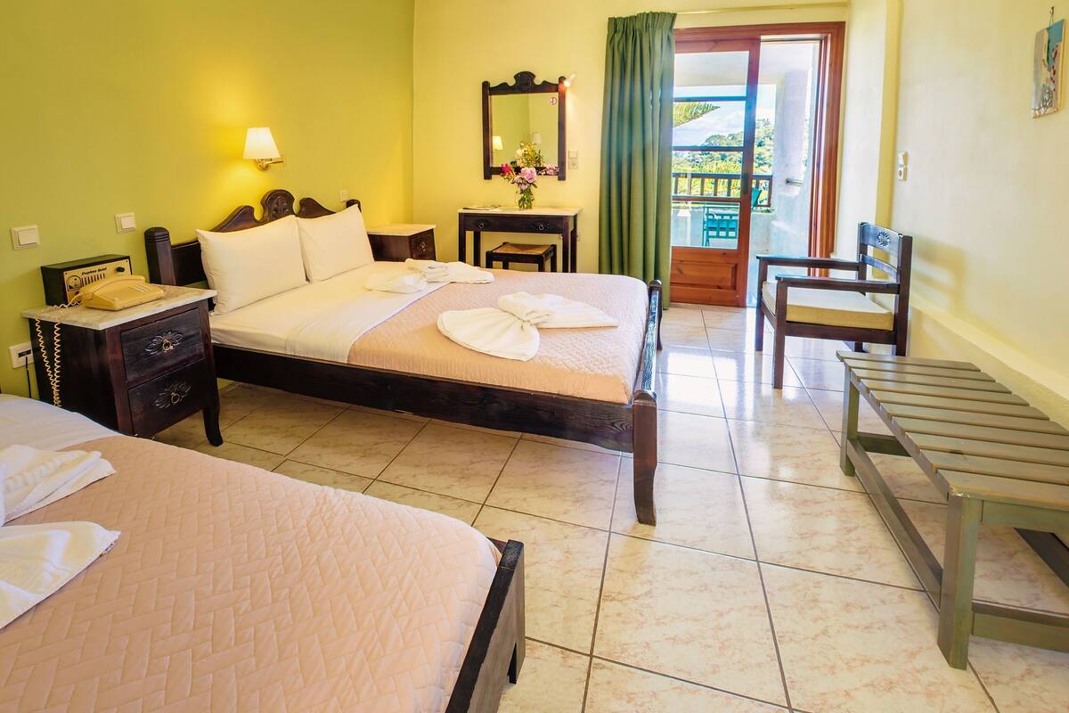 A hotel room with two beds, a wooden bench, a small table, a nightstand with a telephone, a mirror, and a view of the outside through a door.