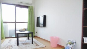 Una habitación pequeña con puerta de vidrio corredera, una mesa con un cesto, un cubo de basura rosa, un cesto verde con revistas y una pared blanca con un televisor montado.