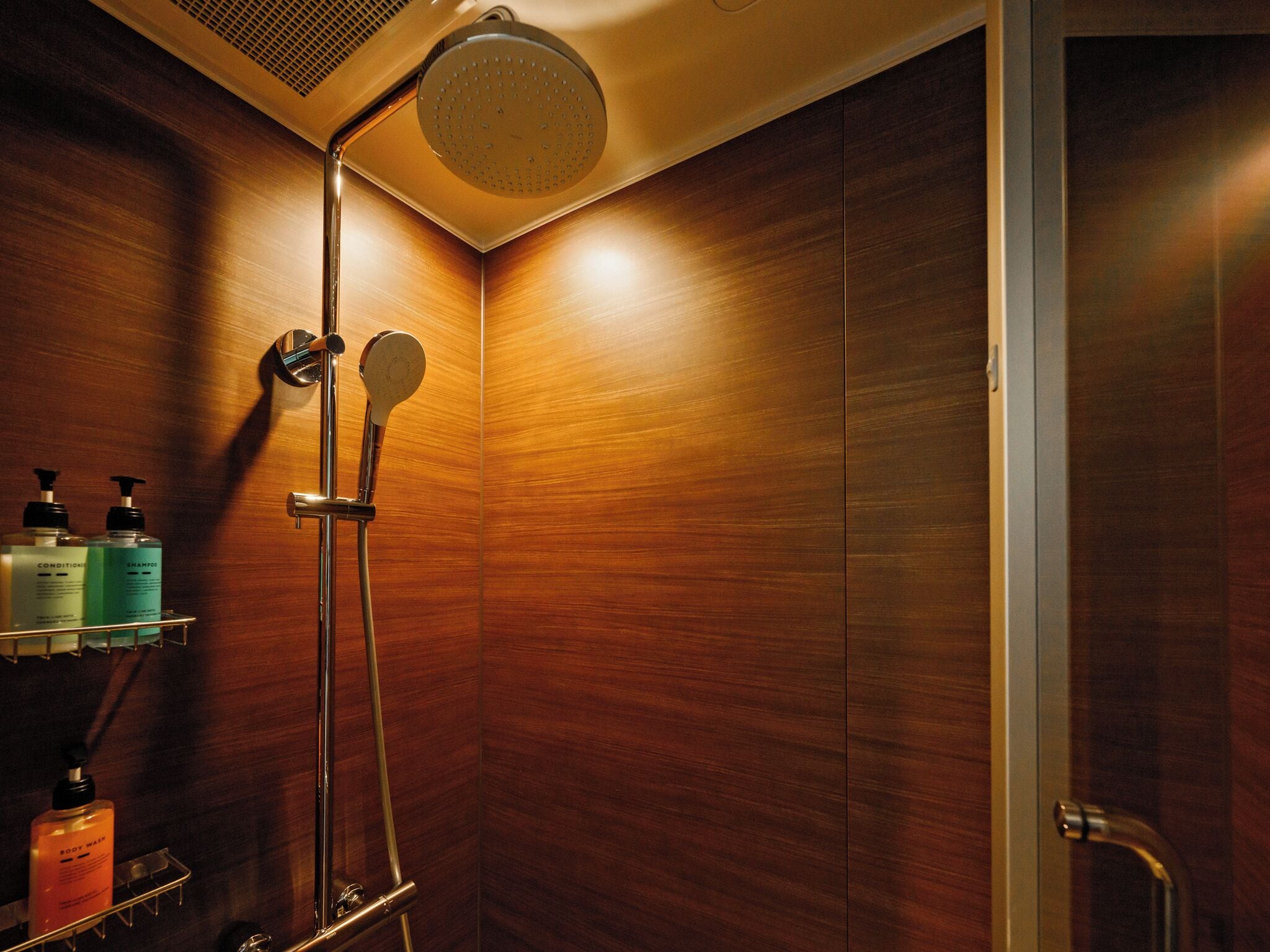 Ducha moderna con cabezal de ducha de lluvia, cabezal de ducha manual y mando a presión en la pared. Hay repisas con artículos de aseo, incluyendo champú y gel de baño.