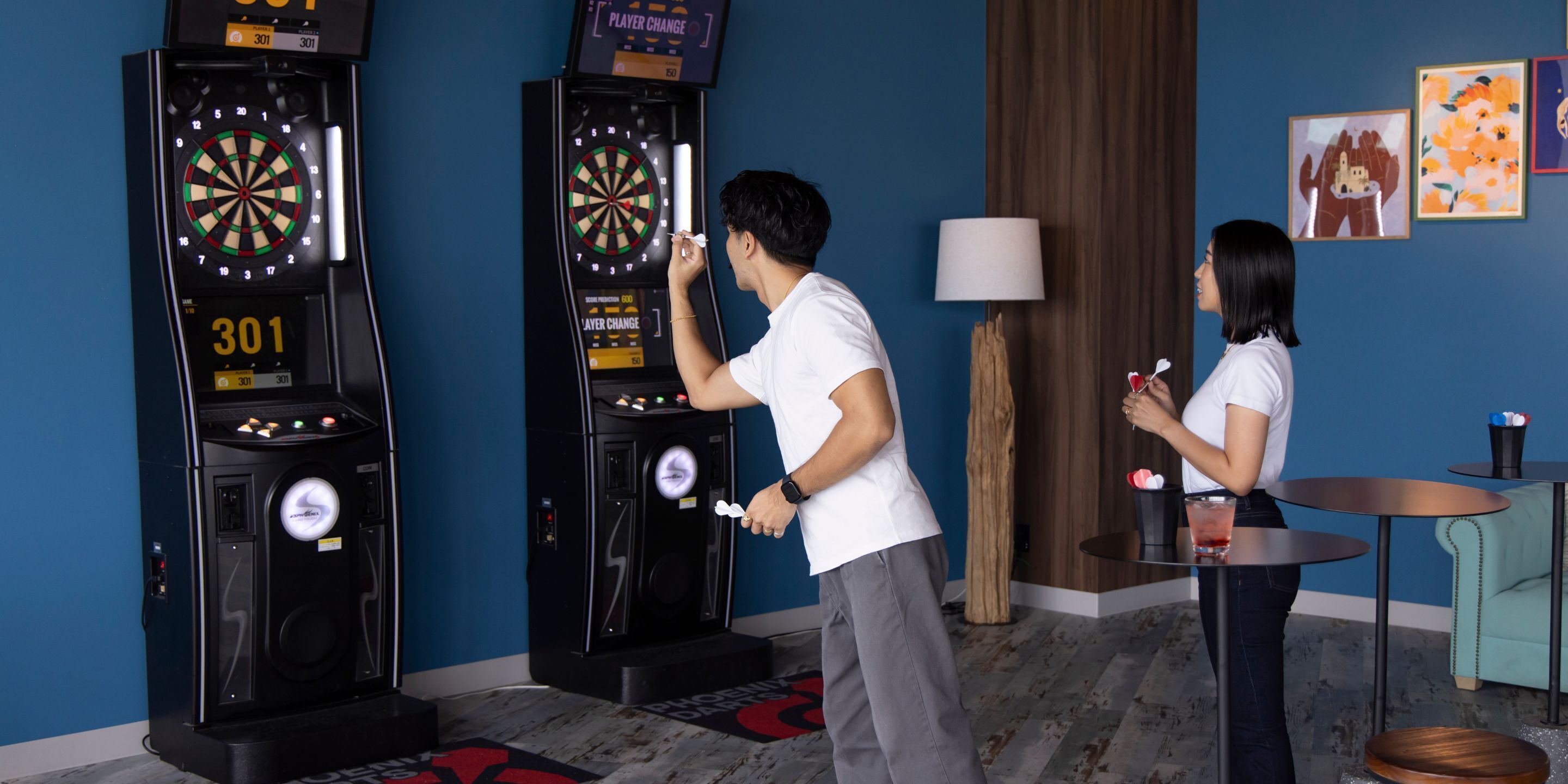 Deux jeux de fléchettes d’arcade dans une pièce avec un mur bleu, une lampe et une petite table sur laquelle se trouve une boisson.