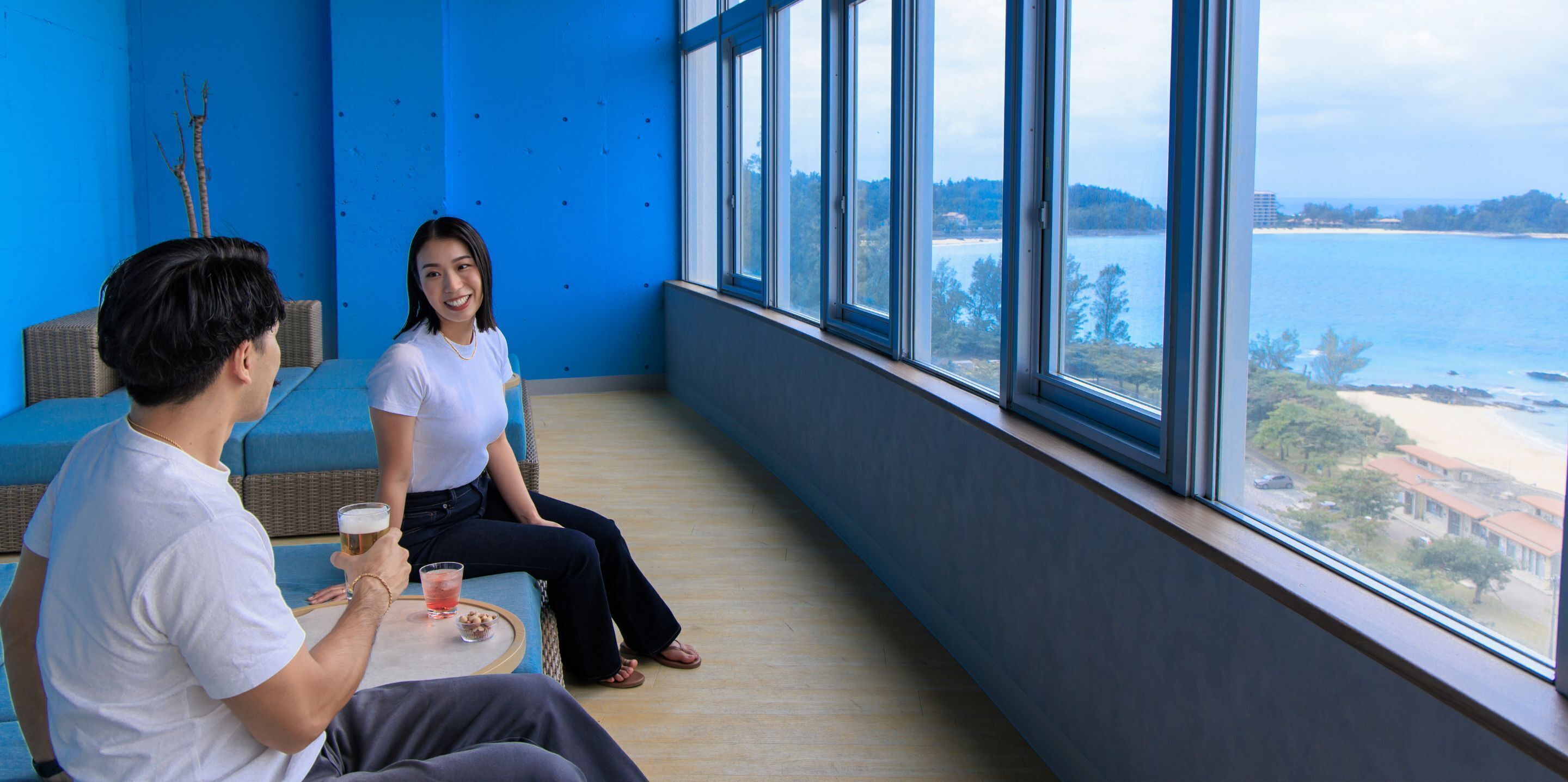 A room with large windows offering a view of the sea and beach. There are two people seated at a table with drinks.