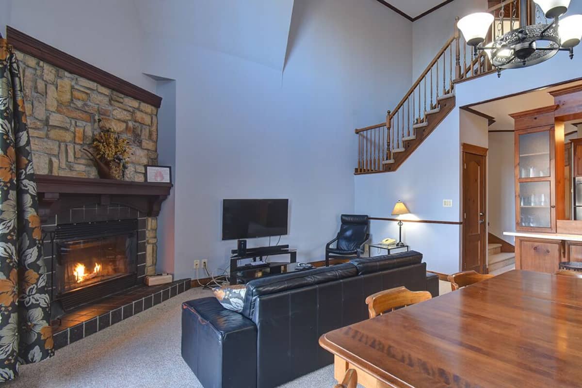 A spacious living room with a stone fireplace, a black leather sofa, a wooden dining table, and a staircase leading to another level.