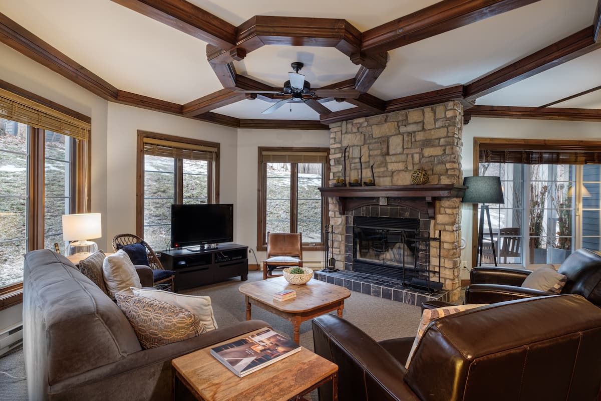 A cozy living room with a stone fireplace, a ceiling fan, and a large window overlooking a snowy landscape.