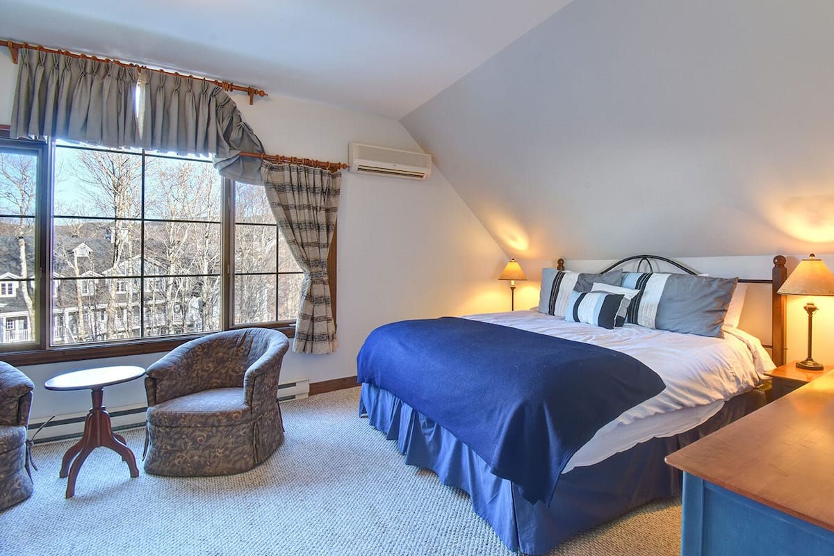 A bedroom with a large bed, two bedside lamps, a chair, a small table, and a view of trees through a window.