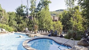 A hot tub surrounded by lounge chairs and a swimming pool, set in a landscaped garden with trees and a view of houses in the background.