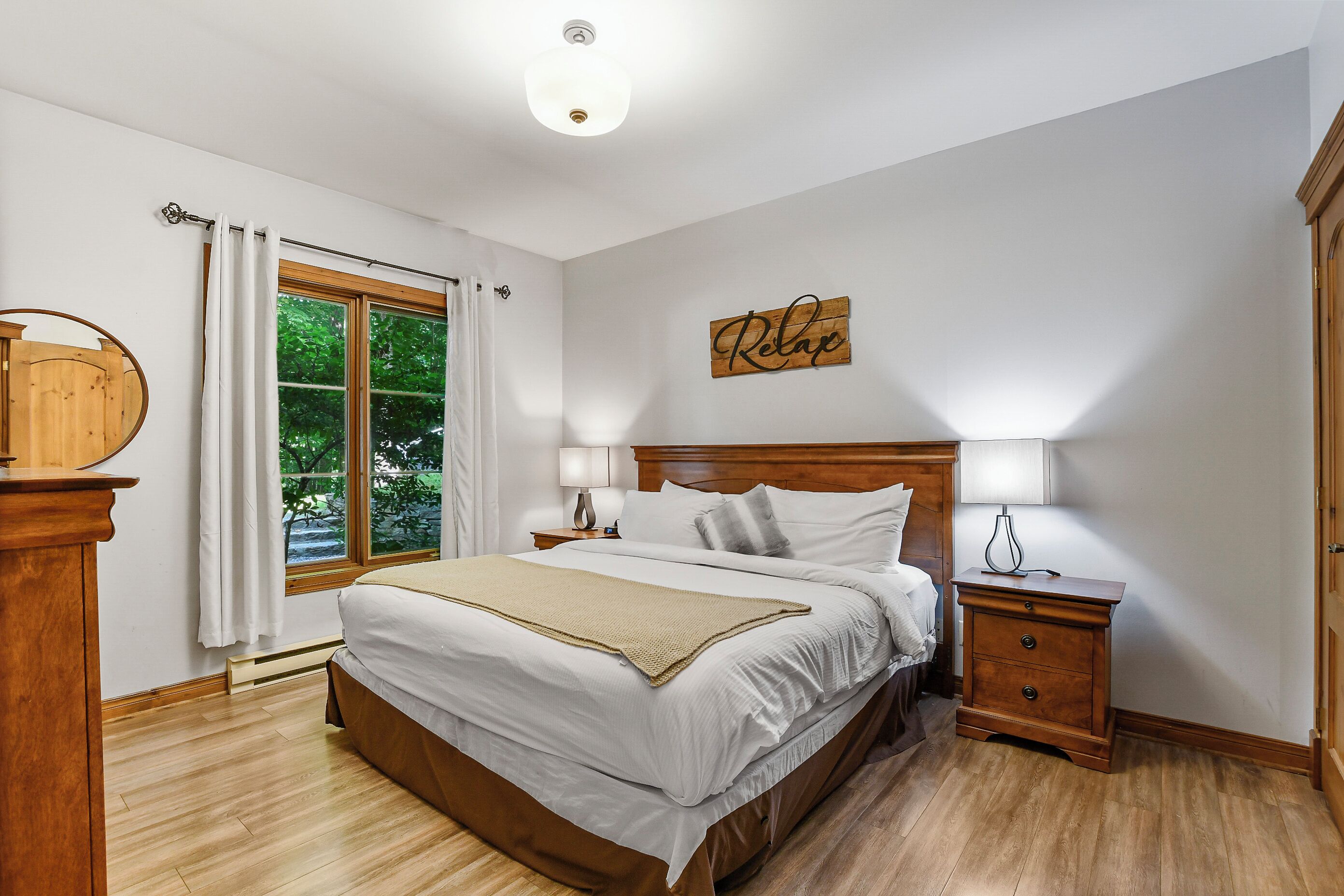 A bedroom with a large bed, wooden headboard, bedside tables with lamps, a wooden dresser, and a window with curtains.