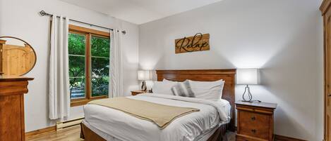 A bedroom with a large bed, wooden headboard, bedside tables with lamps, a wooden dresser, and a window with curtains.