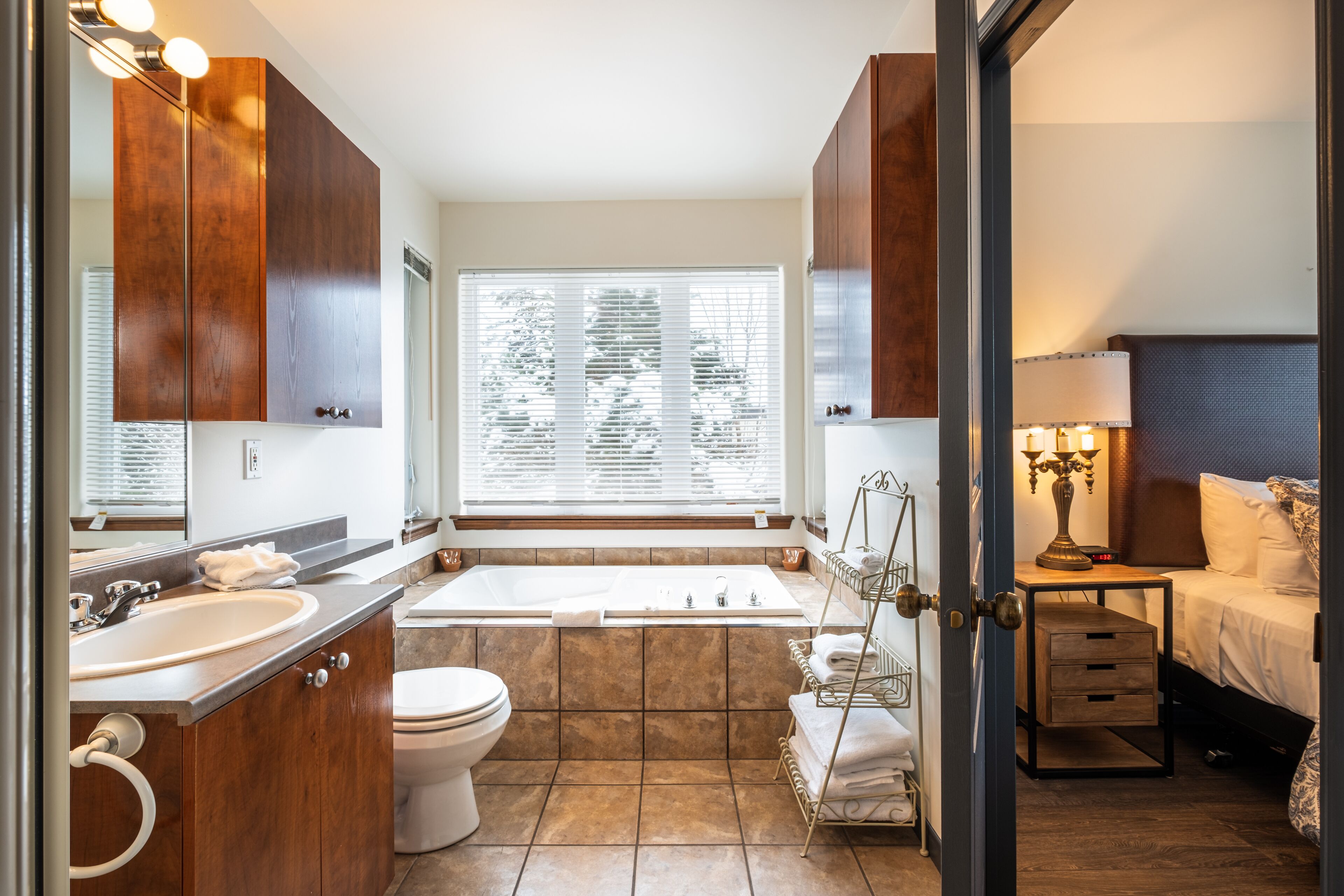 Ein modernes Badezimmer mit Badewanne, Waschbecken und Toilette. Ein Fenster mit Jalousien, ein Handtuchhalter und ein Nachttisch mit Lampe sind vorhanden.