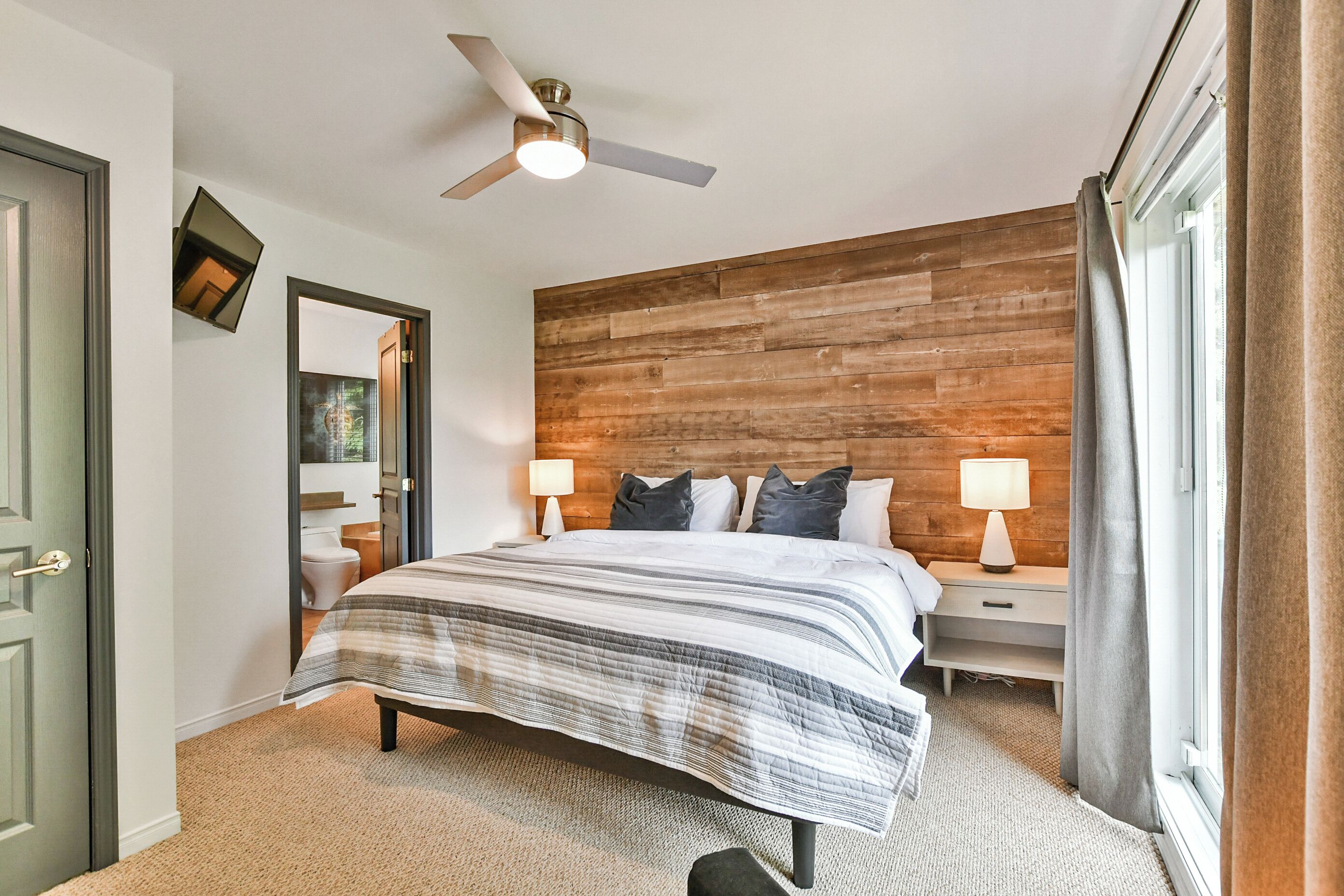 A bedroom with a wooden headboard, a bed with a striped bedspread, two bedside tables with lamps, a ceiling fan, and a view of a bathroom through an open door.