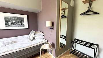 A neatly made bed with a pillow, a small bedside table, a wall-mounted mirror, and a black hanger rack.