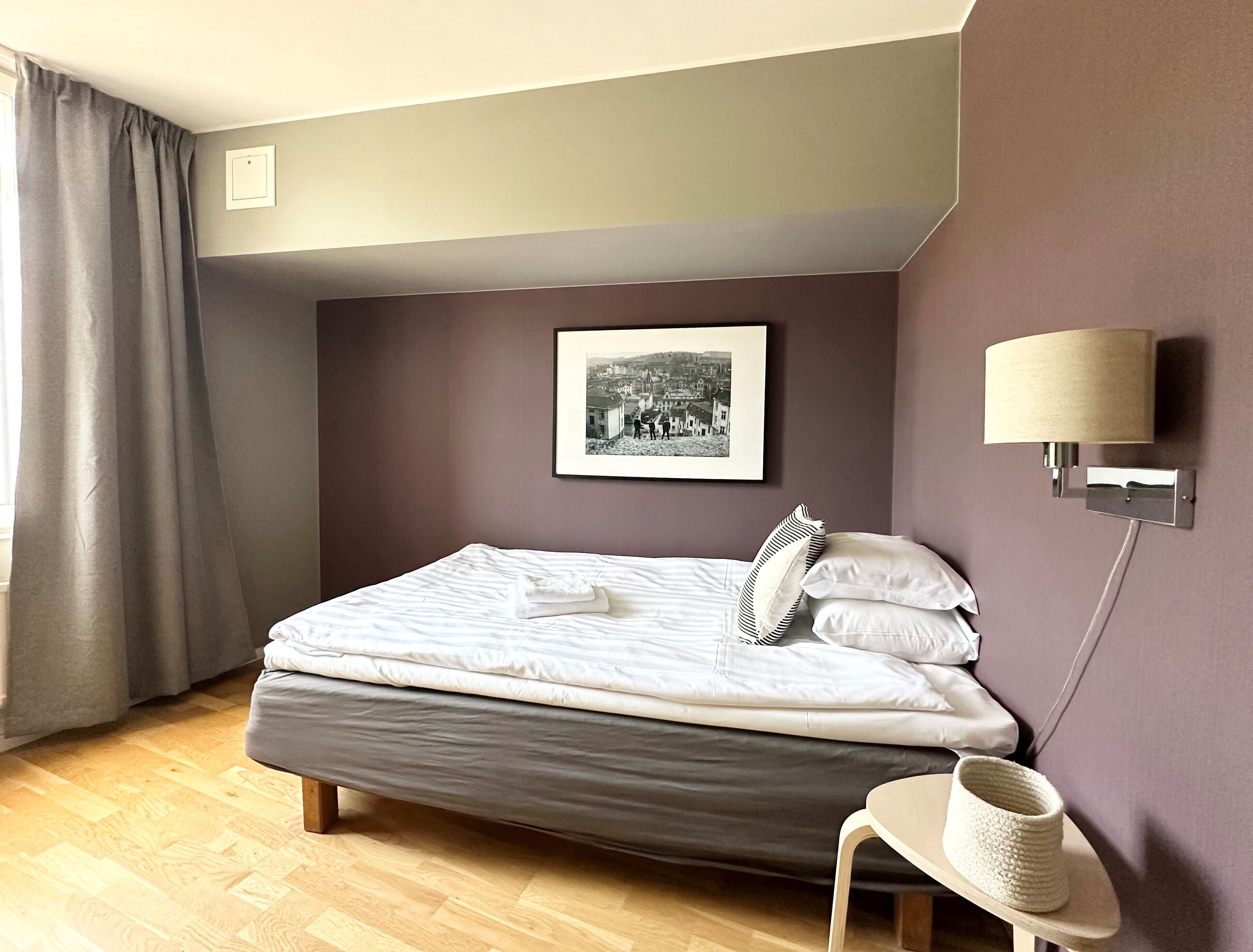 Un lit bien fait dans une pièce au plancher de bois, avec un tableau encadré au mur et une lampe sur la table de chevet.