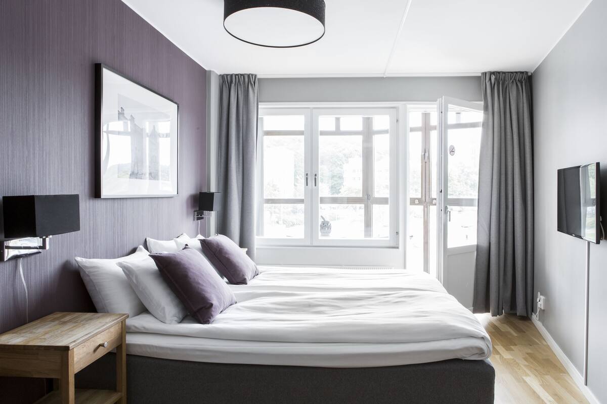 Ein modernes Schlafzimmer mit einem großen Bett, Nachttischen, einem an der Wand befestigten Fernseher und Blick ins Freie durch eine Glastür.