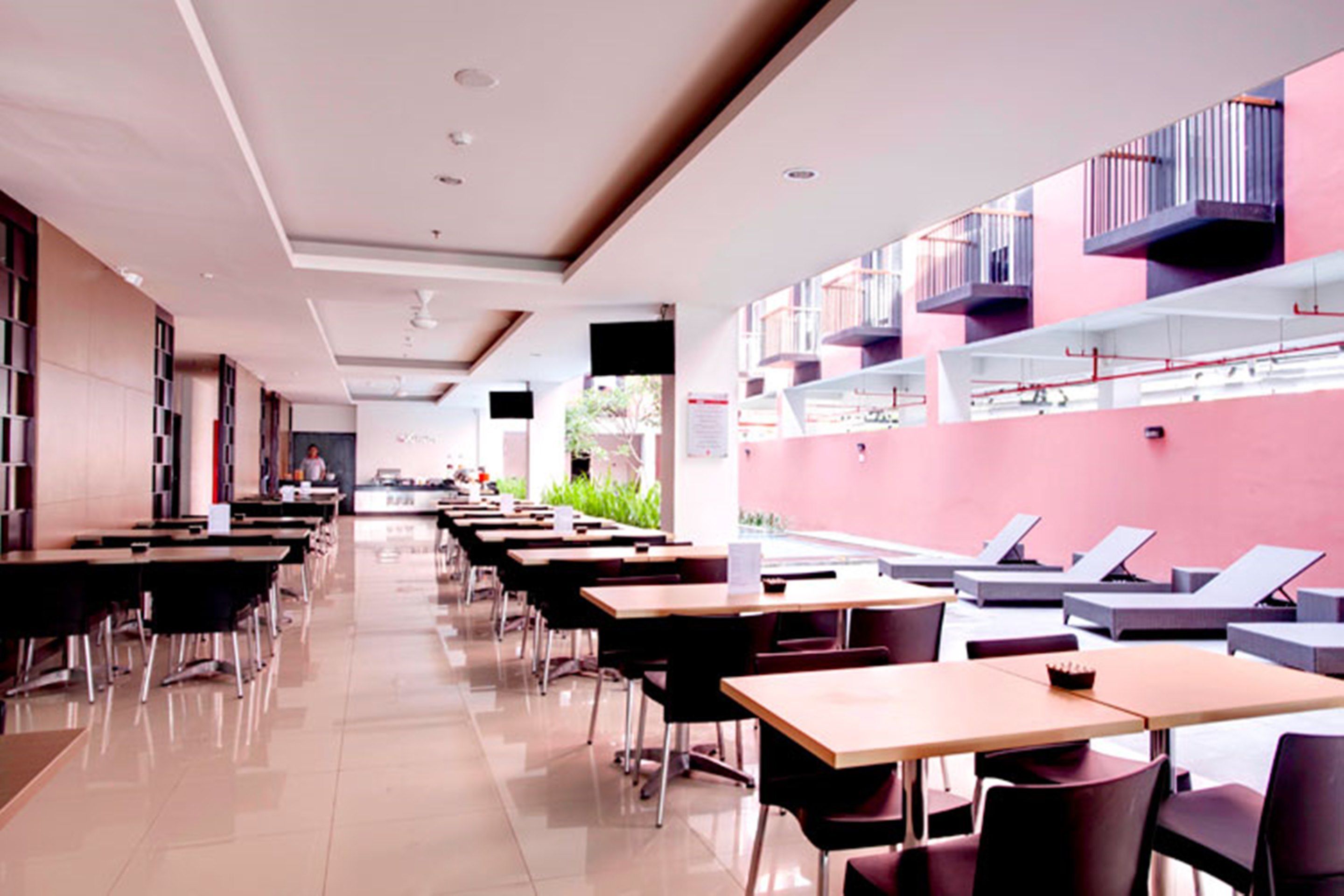 A modern dining area with long tables and chairs, a pink wall with balconies, and a clear view of the outdoors.