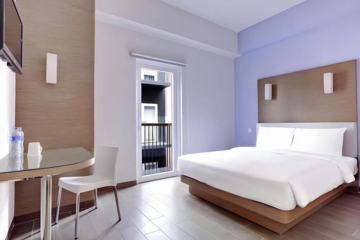 Habitación de hotel con cama, una mesa de vidrio y sillas, una puerta que da a un balcón y un televisor montado en la pared.