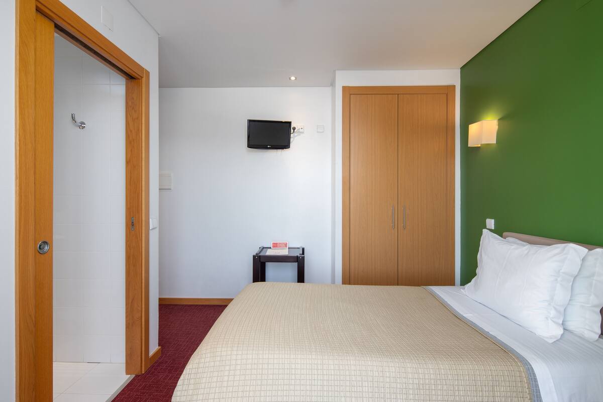 Habitación de hotel con cama, televisor montado en la pared, armario de madera y una pequeña mesa con un cartel.