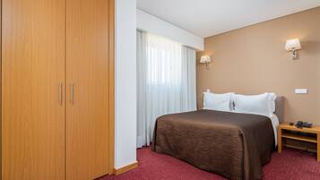 A hotel room with a large wardrobe, a bed with a brown bedspread, white pillows, a bedside table with a lamp, and a red carpet.