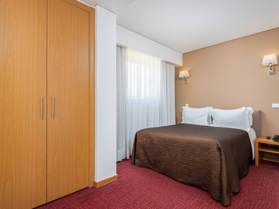 A hotel room with a large wardrobe, a bed with a brown bedspread, white pillows, a bedside table with a lamp, and a red carpet.