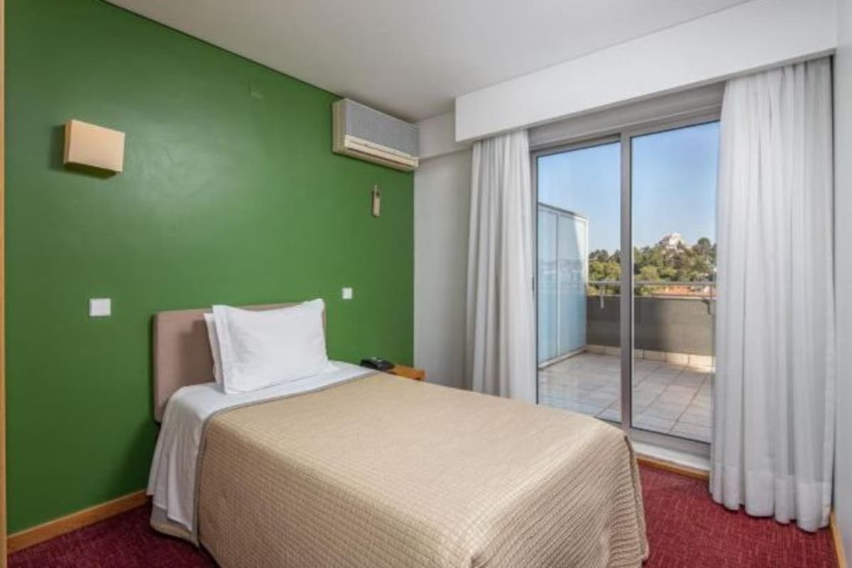 Habitación de hotel con cama, pared verde, equipo de aire acondicionado, puerta corrediza de vidrio a un balcón y vistas al exterior.