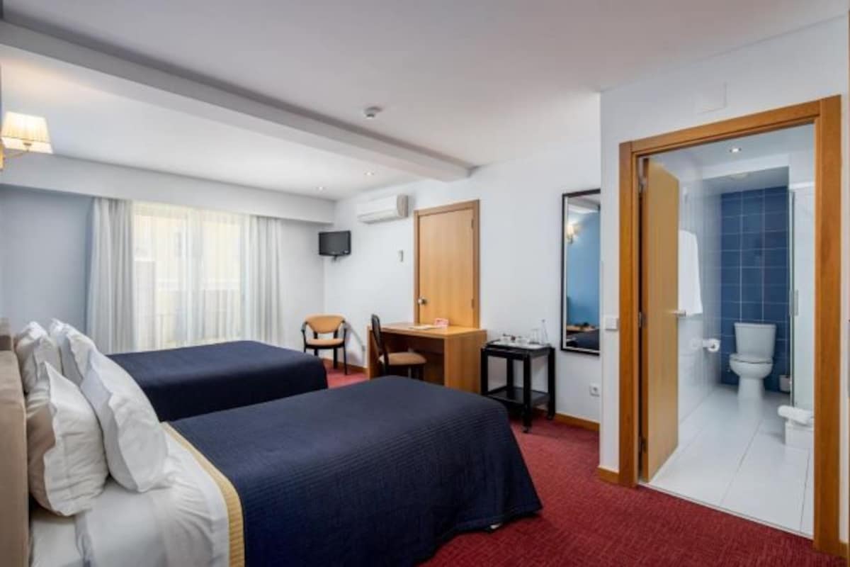 Habitación de hotel con dos camas, un escritorio, una silla, un televisor y un baño visible a través de una puerta abierta.