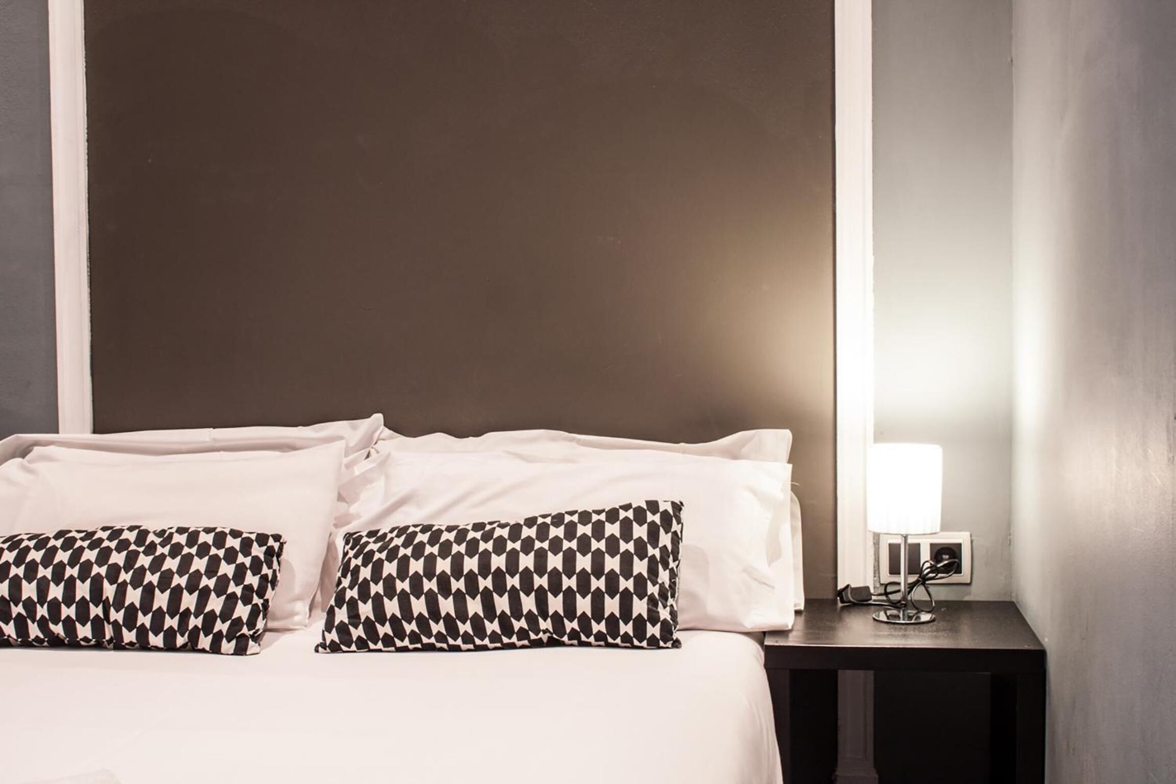 Une chambre d’hôtel avec un lit, deux coussins à motifs, une table de chevet avec une lampe et un luminaire fixé au mur.