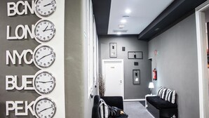 A hotel lobby with a reception desk, a black sofa, a clock with city names, and a fire extinguisher. - Hostal Boutique Khronos (Barcelona)