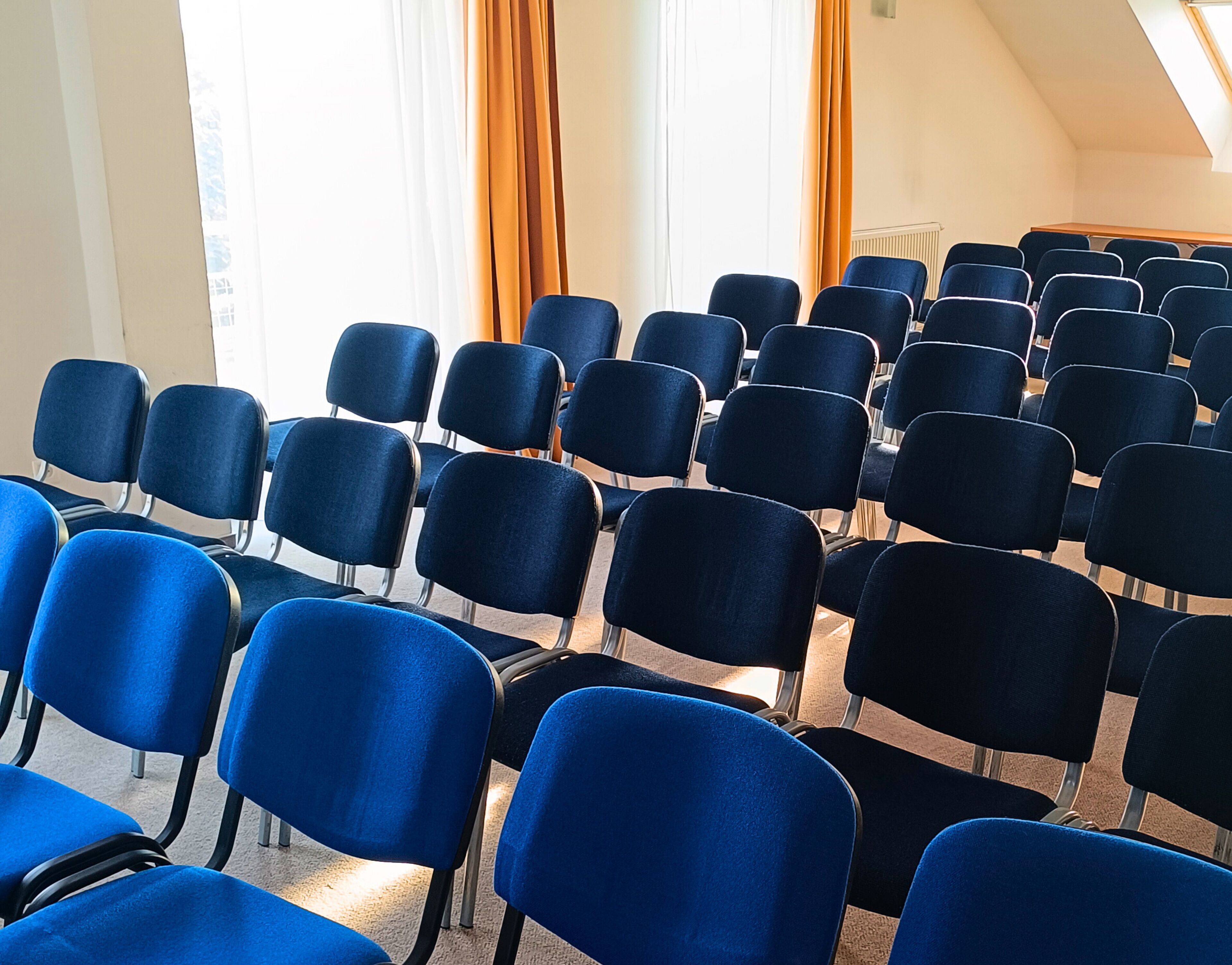 Rijen blauwe stoelen gerangschikt in een congreszaal met een schuine plafond en grote ramen voorzien van gordijnen.