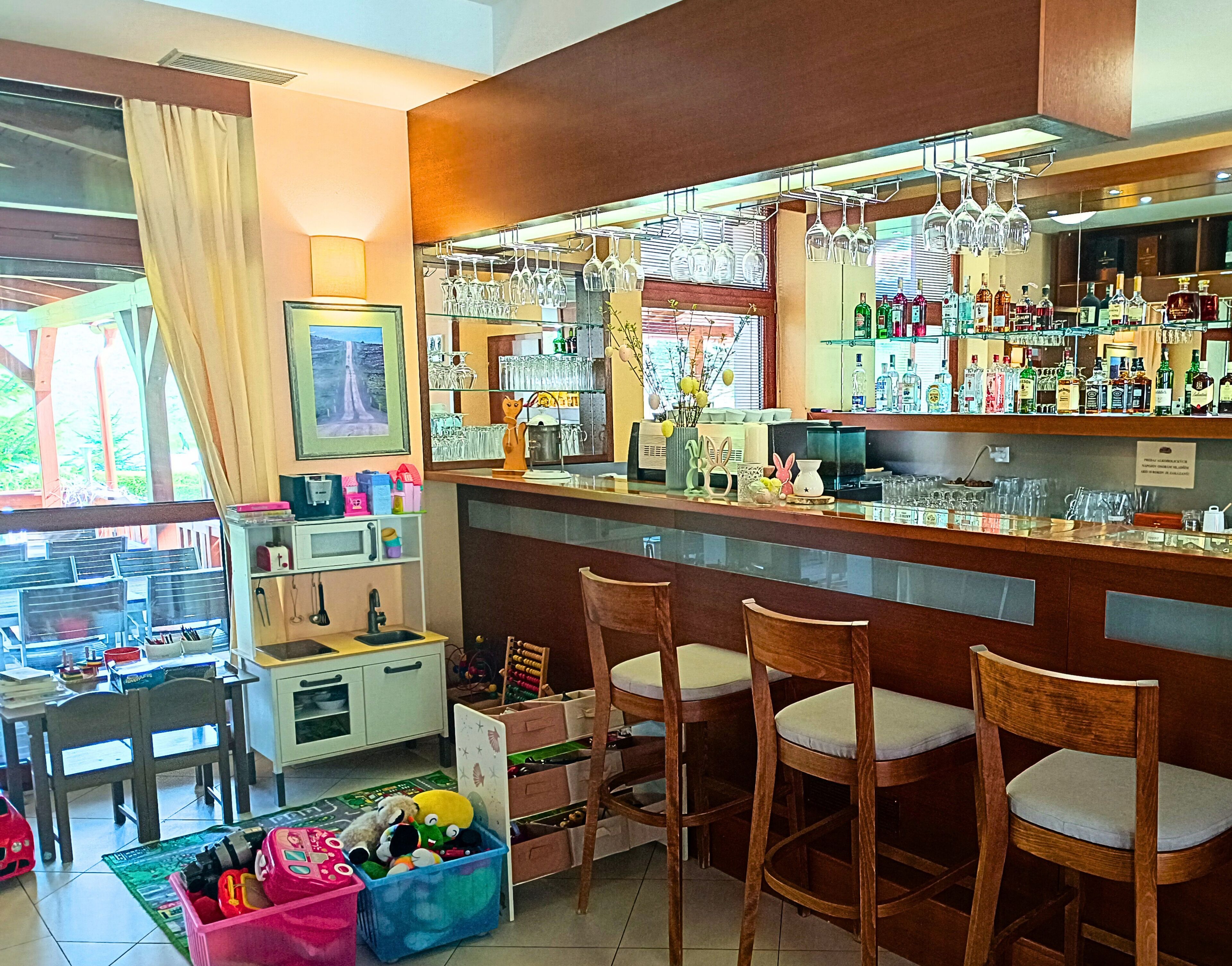 A bar area with wooden chairs, a counter with bottles, and a window with curtains.