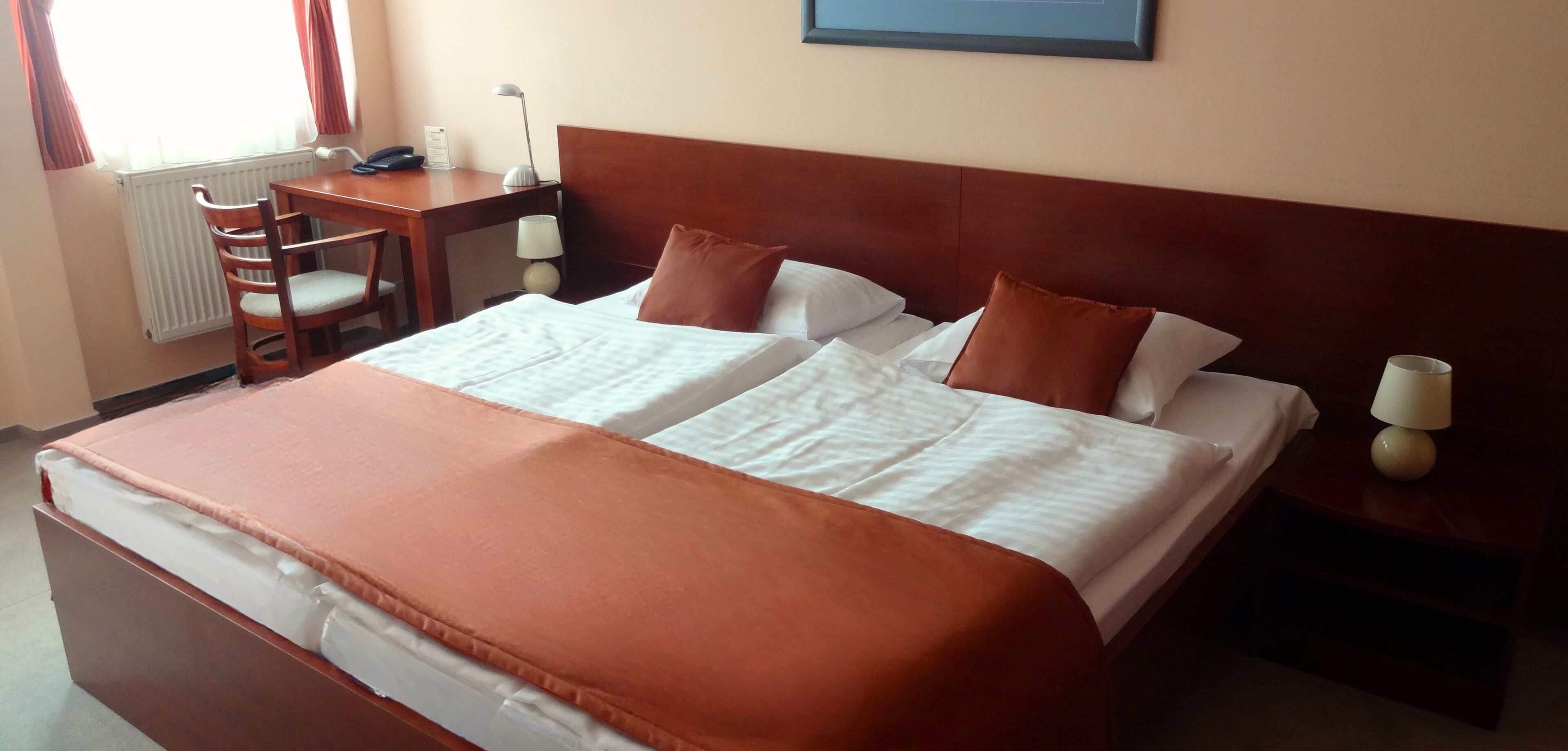 Cama bem arrumada com colcha e travesseiros marrons, mesa de cabeceira de madeira com abajur, uma cadeira de madeira e escrivaninha com abajur e telefone.