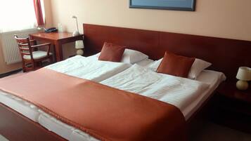 Cama bem arrumada com colcha e travesseiros marrons, mesa de cabeceira de madeira com abajur, uma cadeira de madeira e escrivaninha com abajur e telefone.