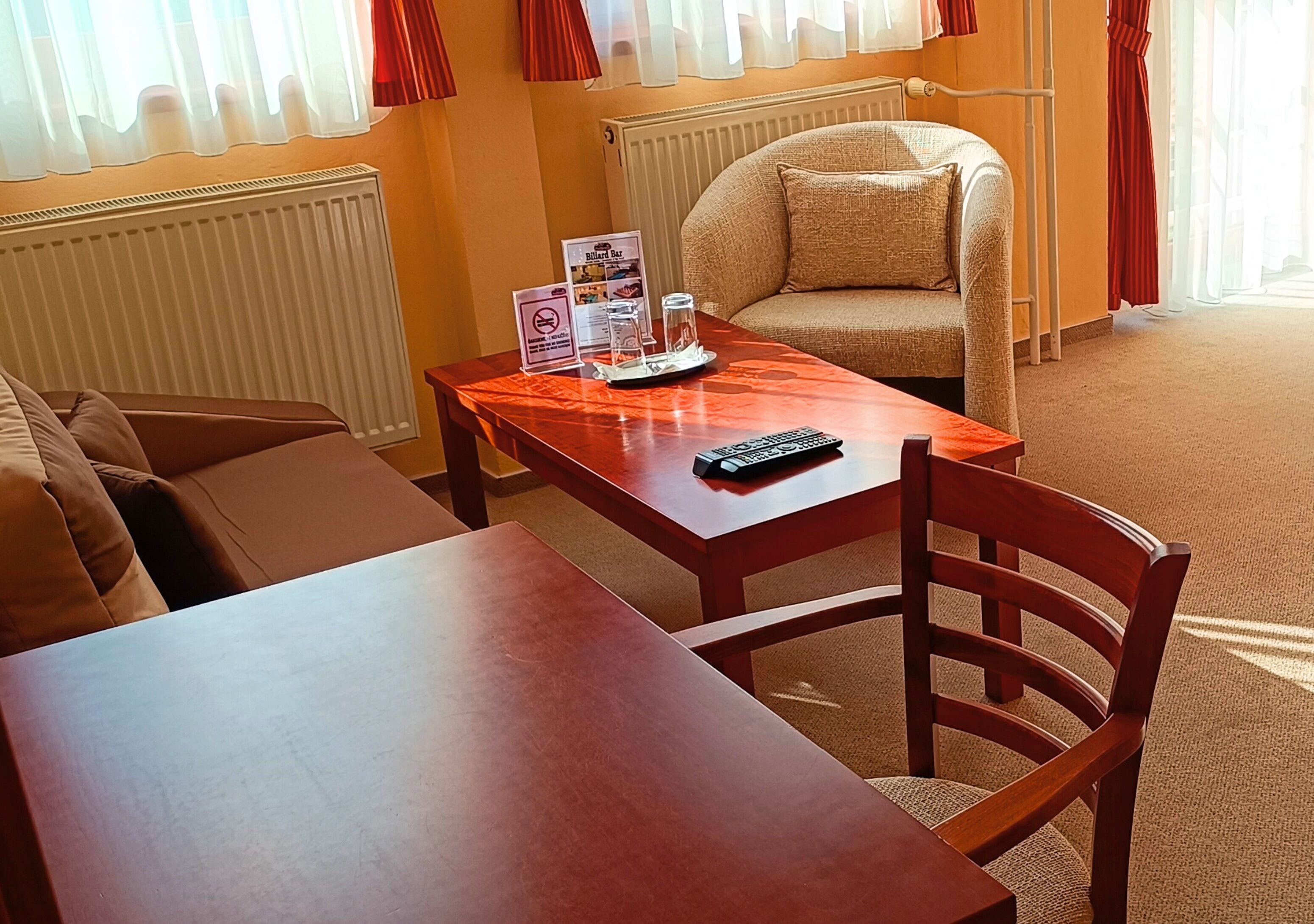 Una habitación de hotel con una mesa de madera, una silla, un sofá, un radiador y una ventana con cortinas.