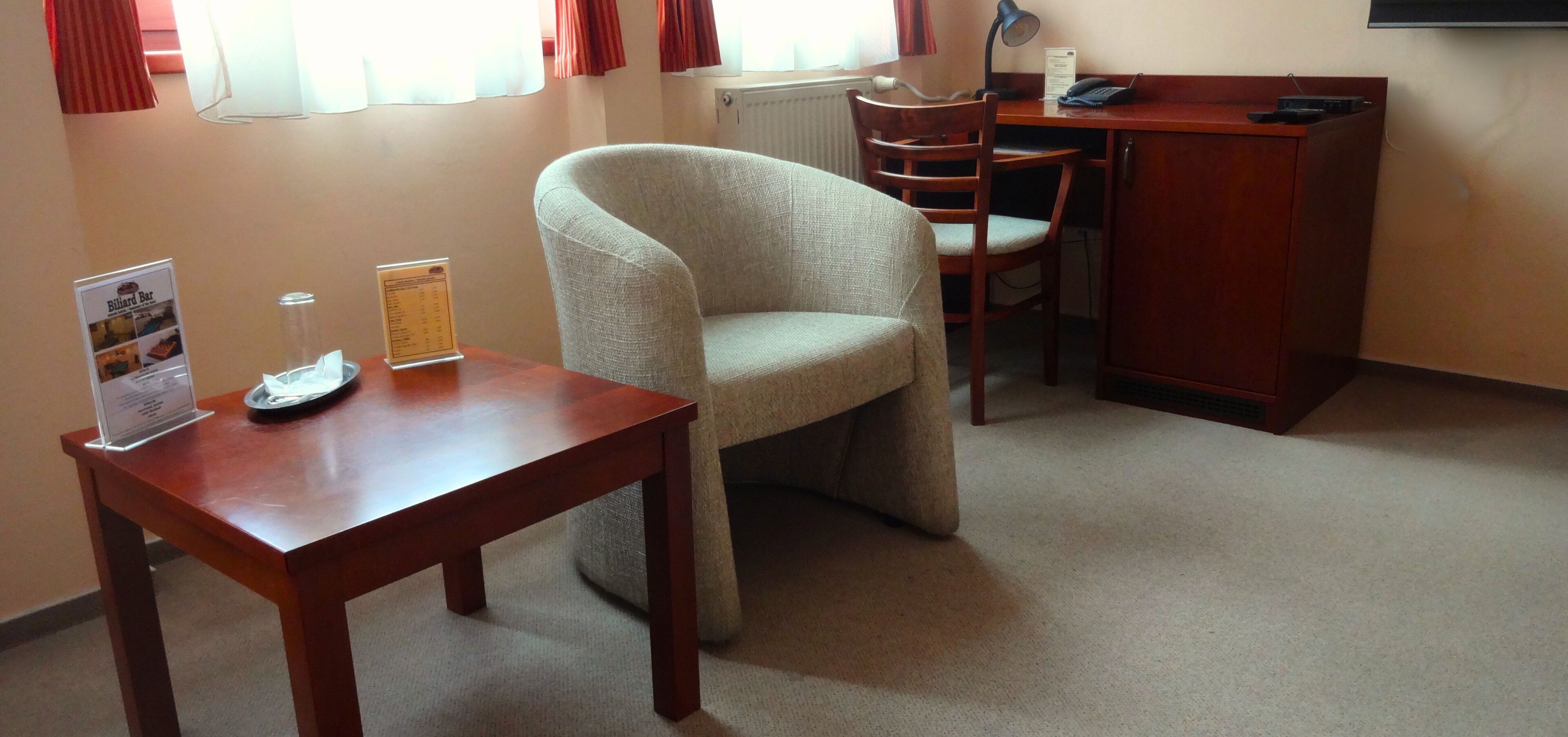 Een hotelkamer met een houten bureau, een comfortabele fauteuil, een houten stoel en een bijzettafeltje met een lamp en een menukaart.