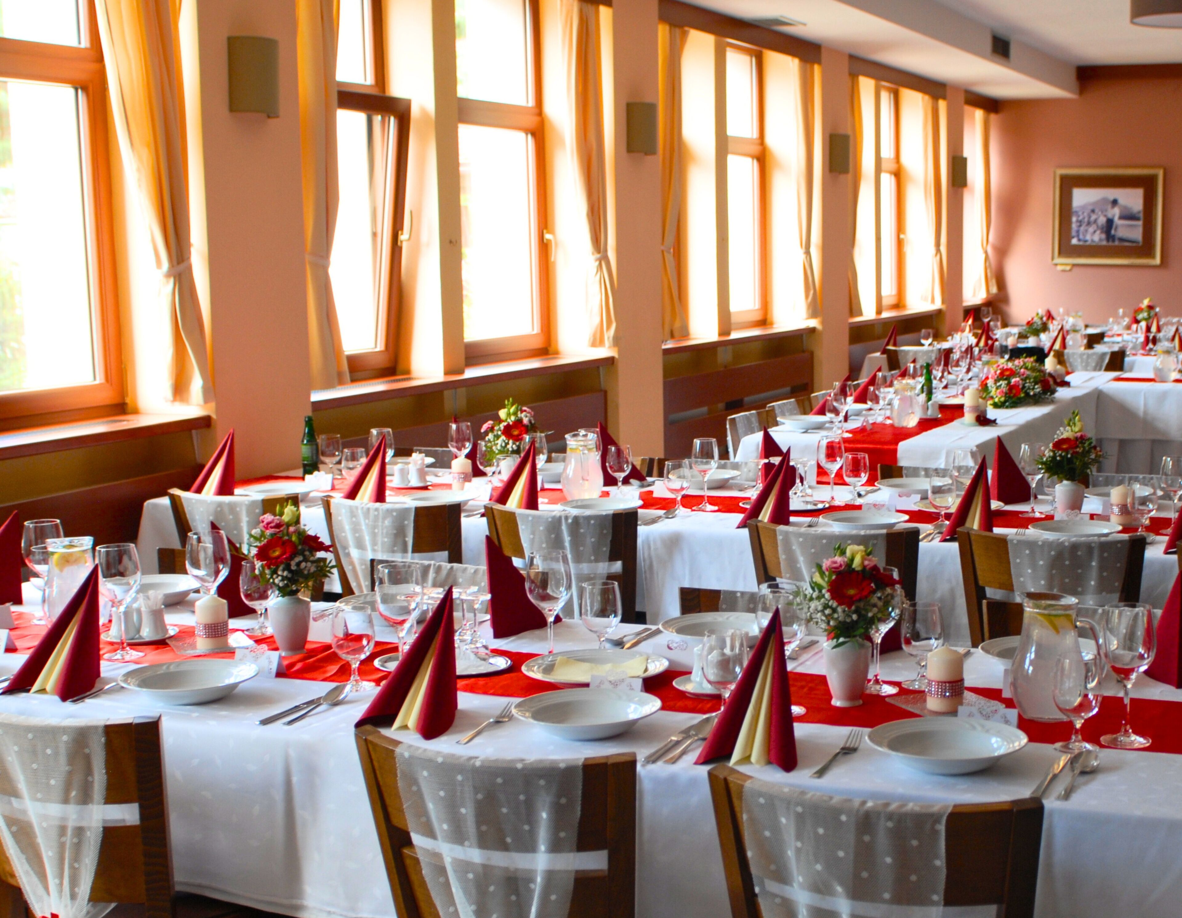 Un salón de banquetes con varias mesas dispuestas para comer, con manteles blancos, servilletas rojas y centros de mesa florales.