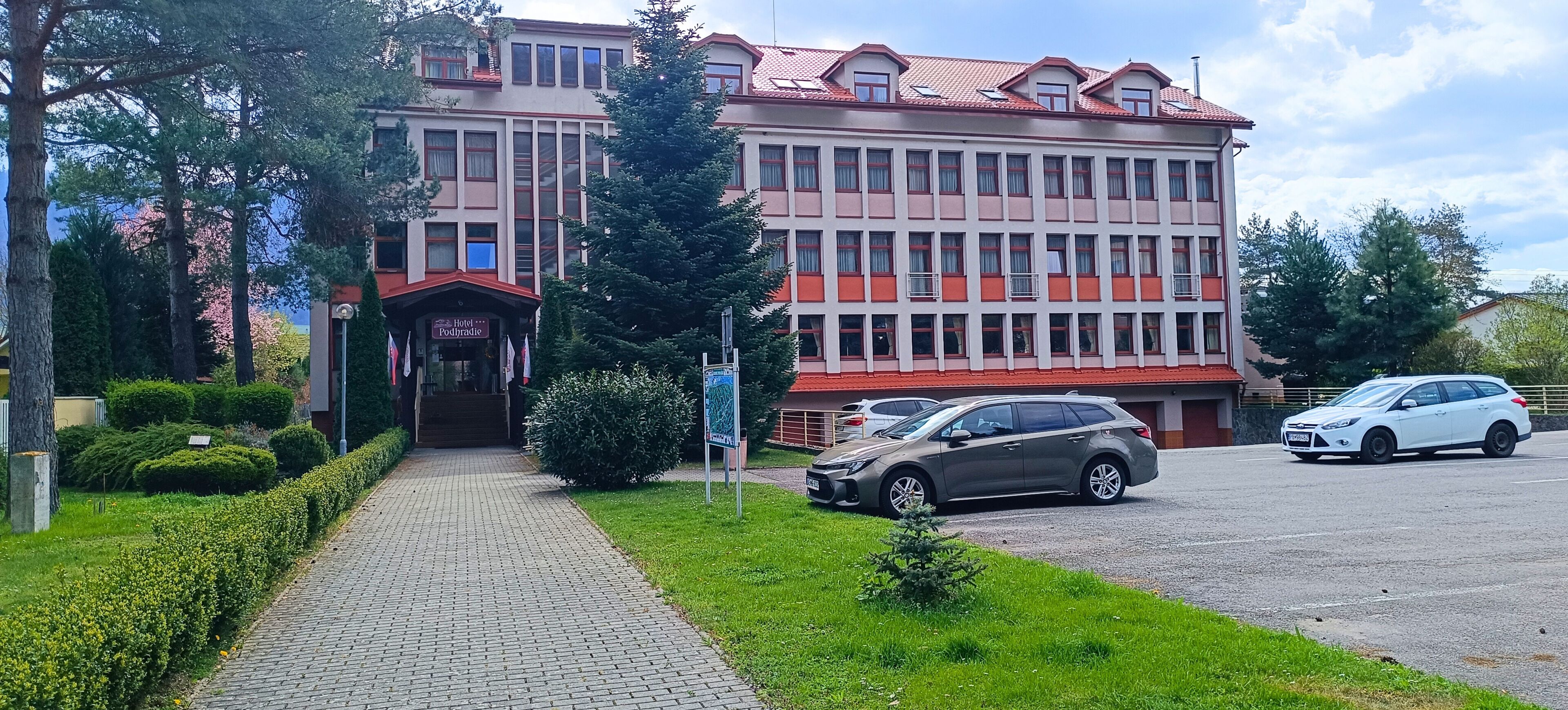 A multi-story hotel with a red and white facade, a paved driveway, and a parking area with cars.