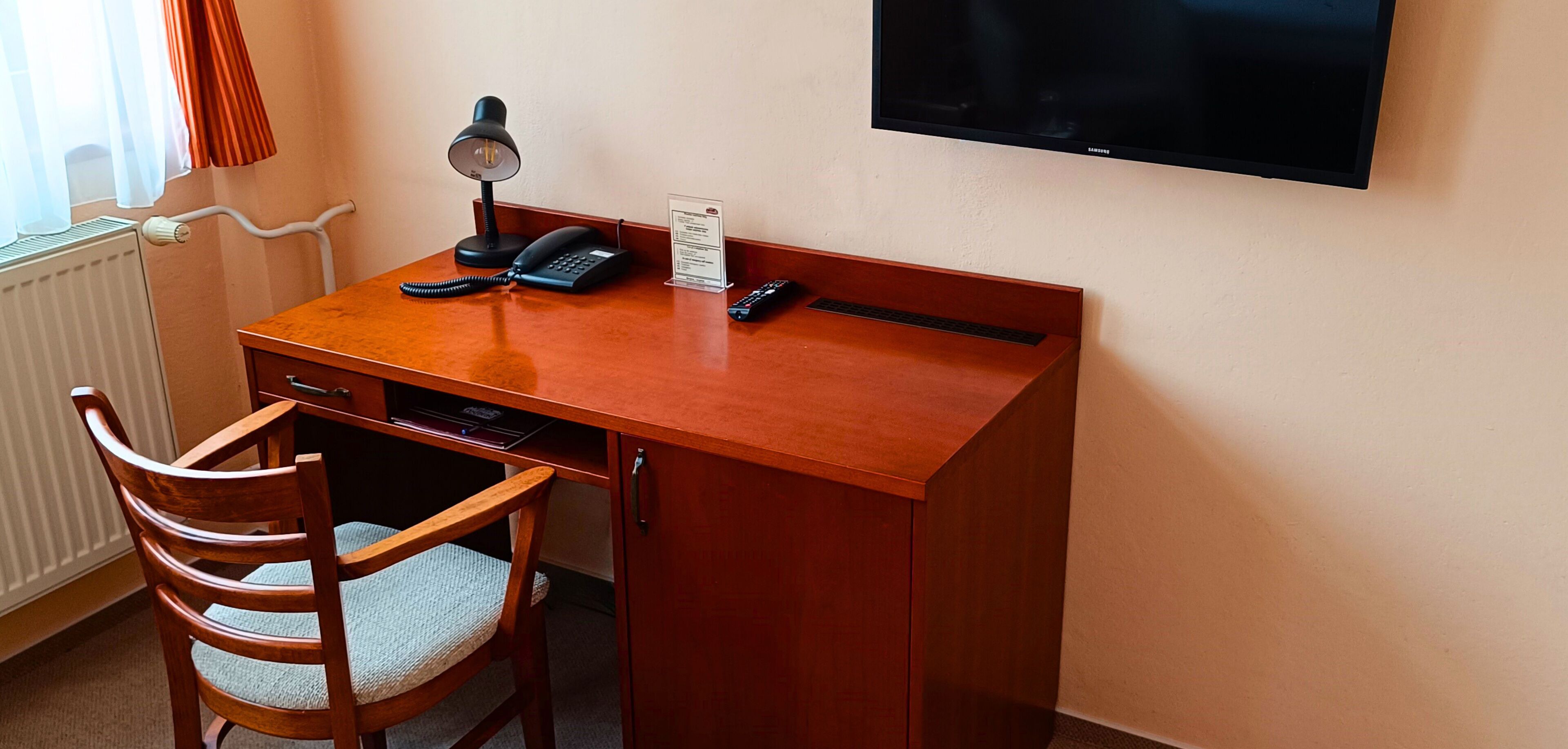 A hotel room with a wooden desk, a telephone, a remote control, a chair, a radiator, and a television mounted on the wall.