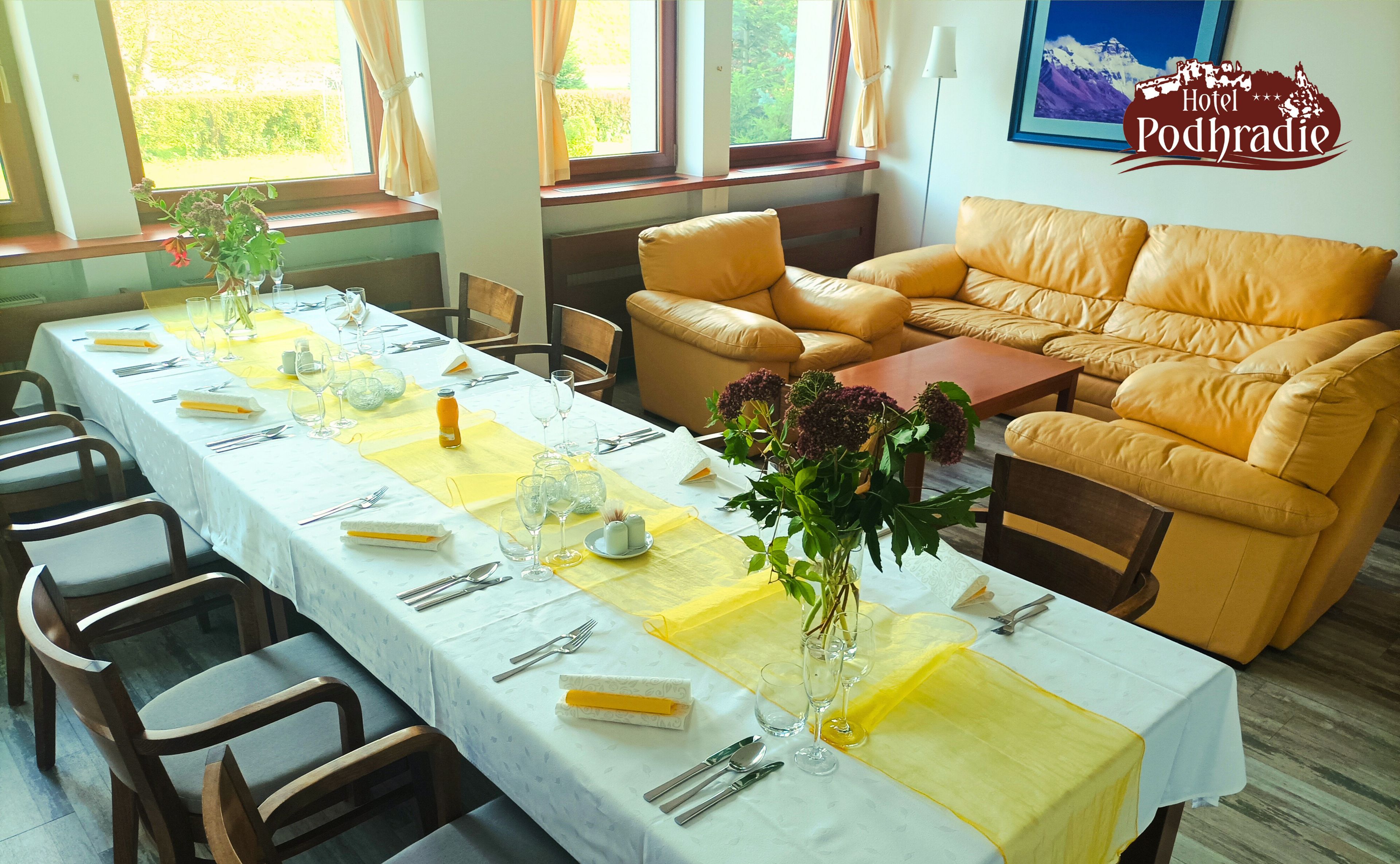 A long dining table set with yellow tablecloths, white tablecloths, and a vase of flowers. There is a sofa and a lamp in the background.